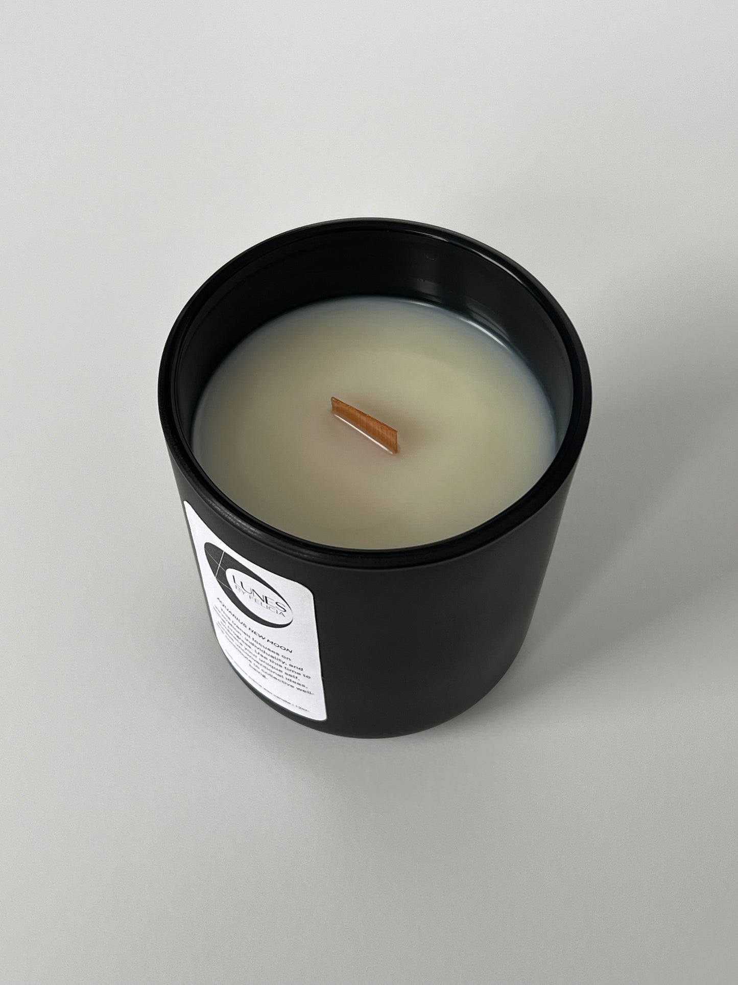 A top view of a matte black glass candle vessel with a white label on the front, featuring the Lunes by Felicia black crescent moon logo. A single flat wooden wick centered in white coconut-apricot wax. The matte black candle vessel is set against a white background.