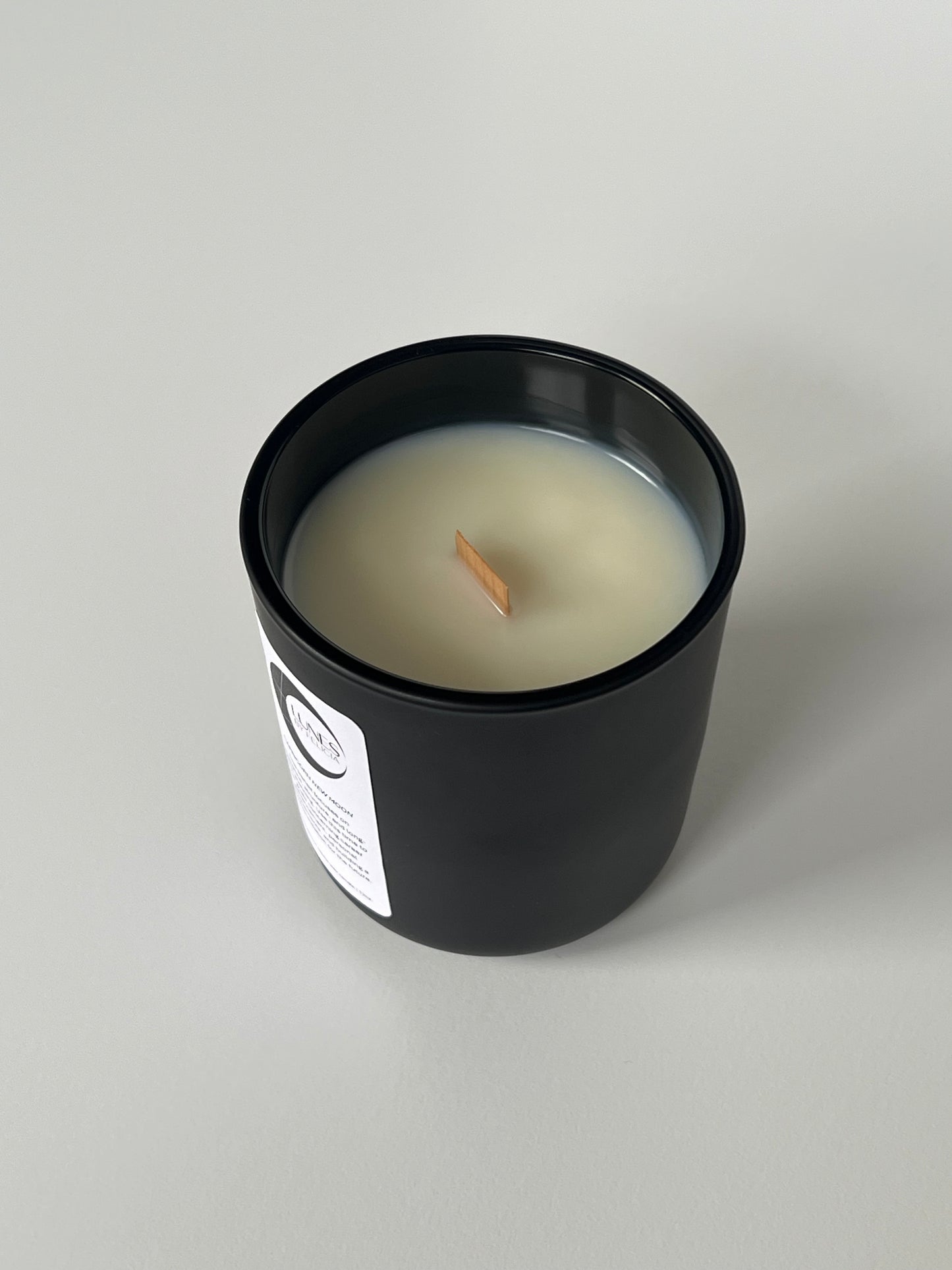 A top view of a matte black glass candle vessel with a white label on the front, featuring the Lunes by Felicia black crescent moon logo. A single flat wooden wick centered in white coconut-apricot wax. The matte black candle vessel is set against a white background.