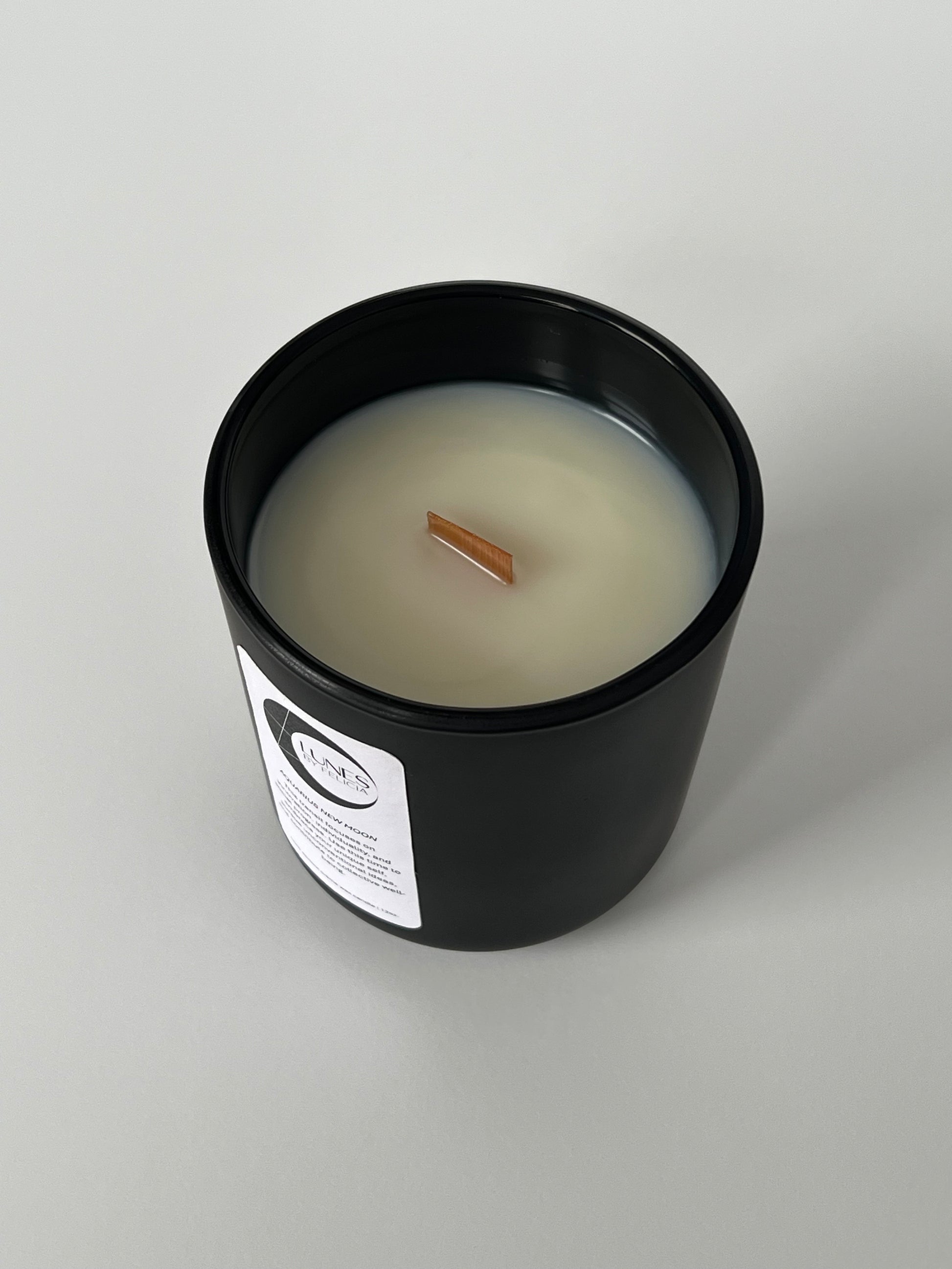 A top view of a matte black glass candle vessel with a white label on the front, featuring the Lunes by Felicia black crescent moon logo. A single flat wooden wick centered in white coconut-apricot wax. The matte black candle vessel is set against a white background.