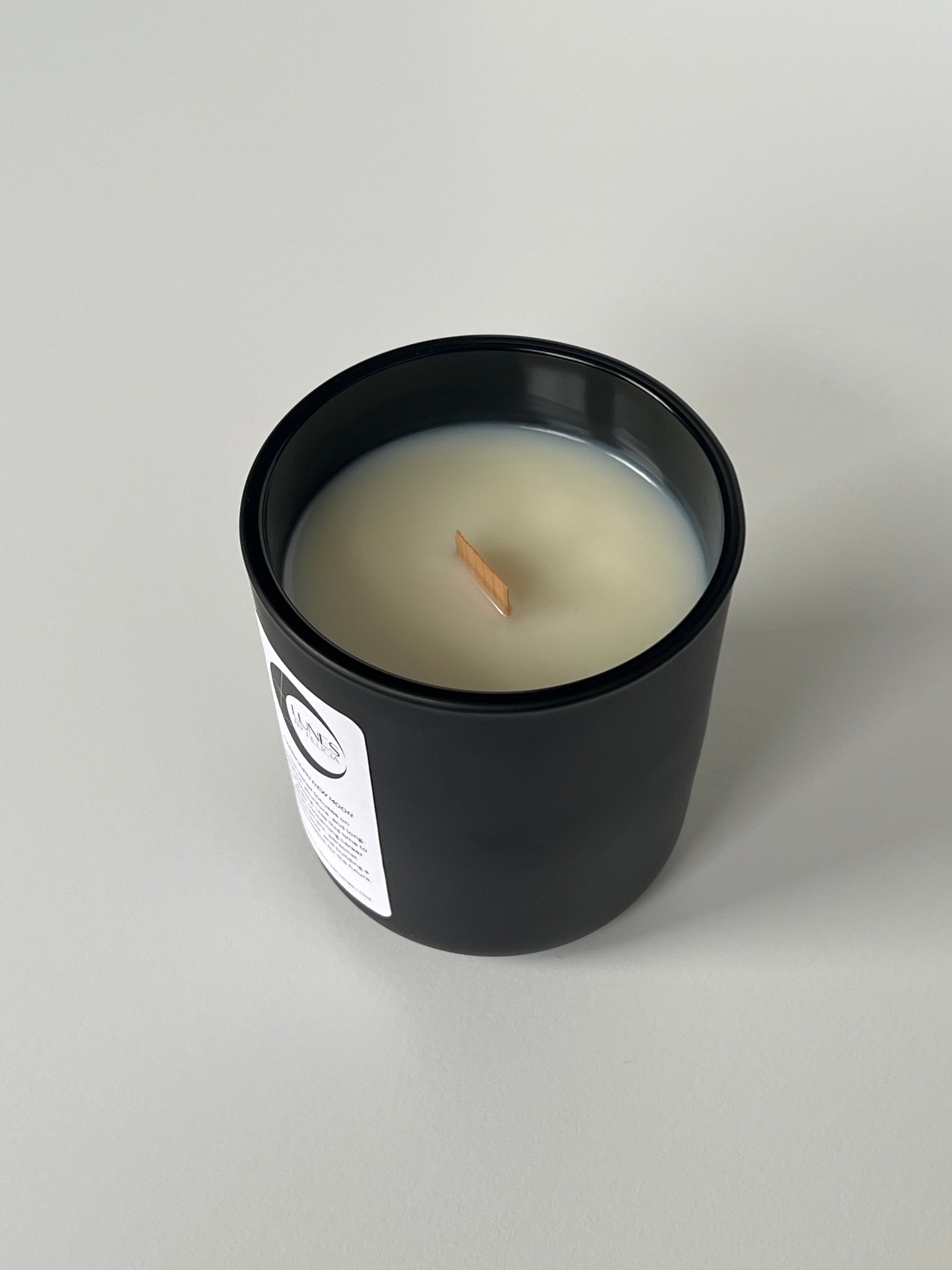 A top view of a matte black glass candle vessel with a white label on the front, featuring the Lunes by Felicia black crescent moon logo. A single flat wooden wick centered in white coconut-apricot wax. The matte black candle vessel is set against a white background.
